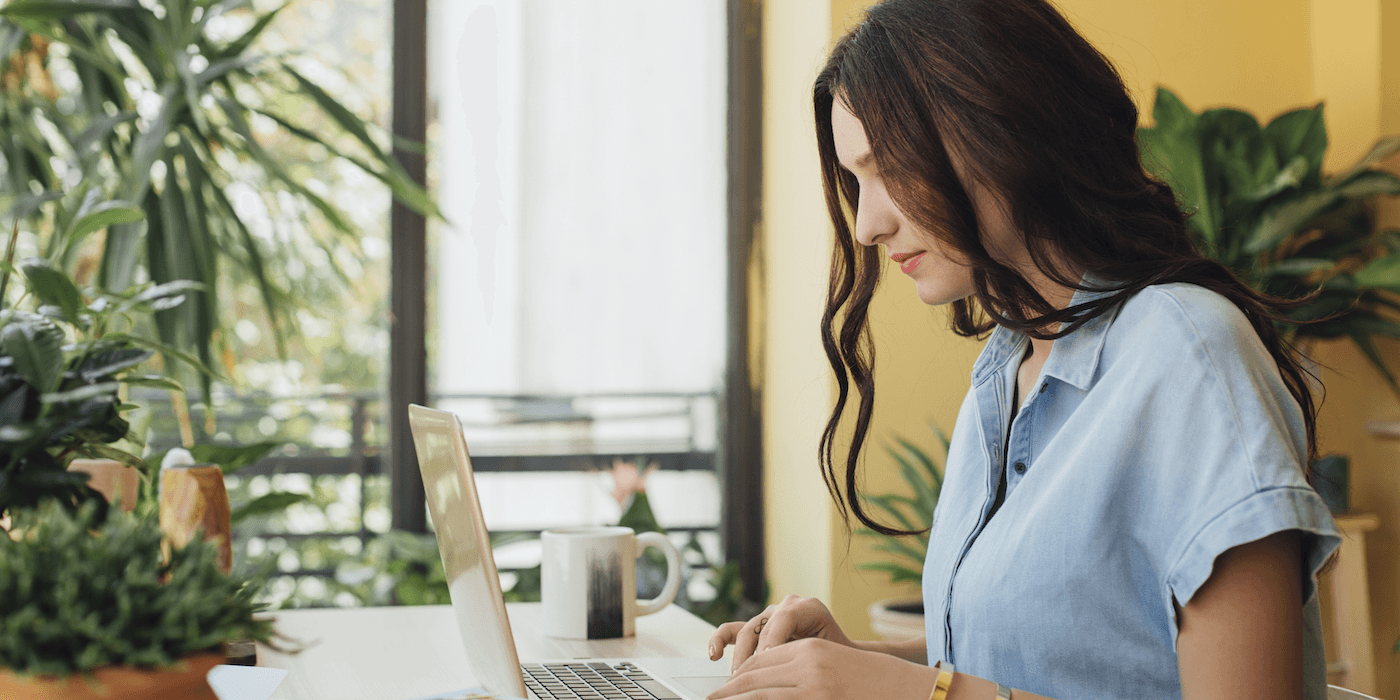 person sitting at a table typing on laptop with a coffee mug and plants nearby