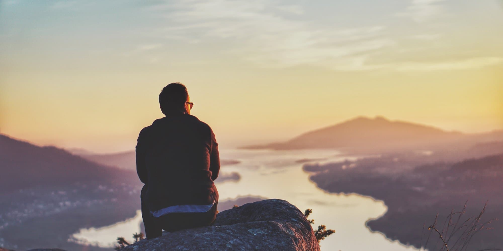 person sitting on a rock at the top of a mountain overlooking a valley with a stream running through it against a yellow, orange, and blue sunset